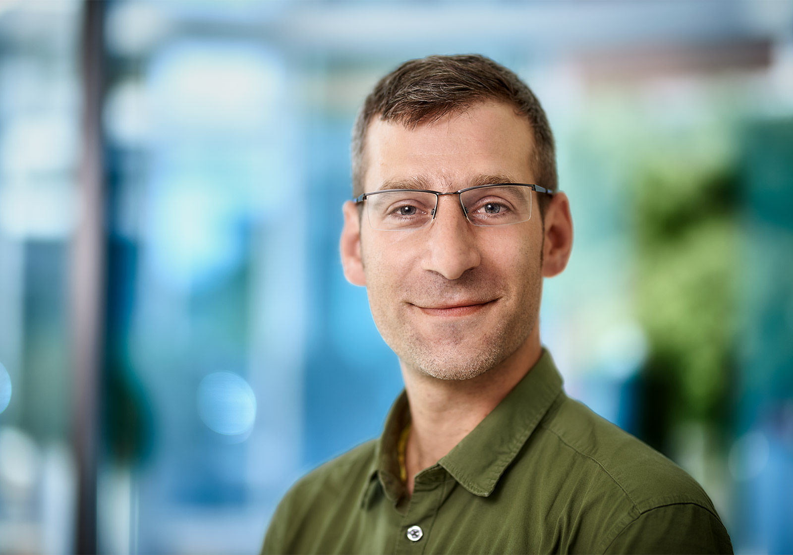 Jakob Rietzler | Rietzler Gruppe Es handelt sich um eine Nahaufnahme von Geschäftsführer Jakob Rietzler. Er trägt ein grünes Hemd, eine Brille und kurze braune Haare. Er lächelt in die Kamera. Der Hintergrund ist unscharf.