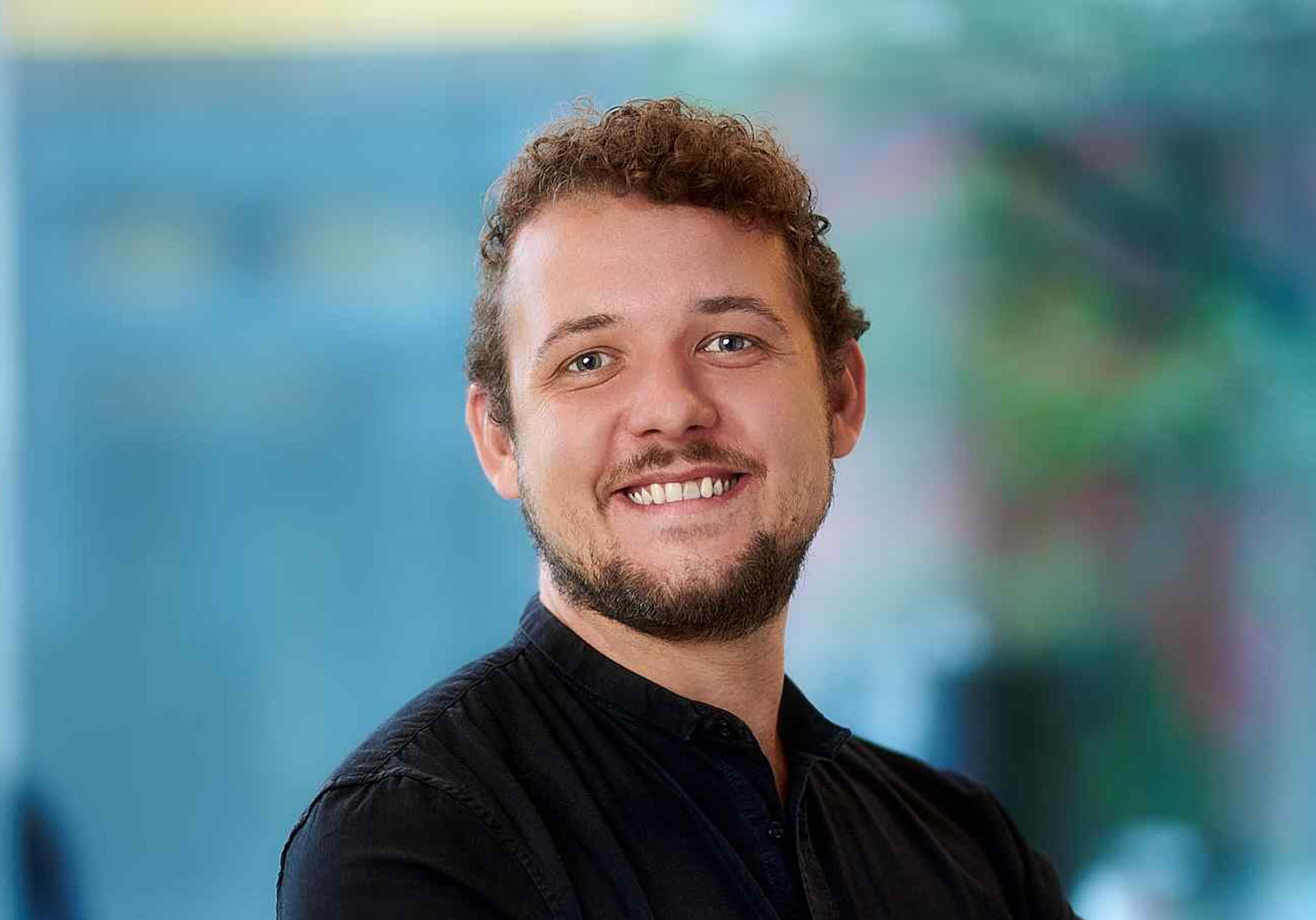 Philipp Samland | Rietzler Gruppe Es handelt sich um eine Nahaufnahme von Geschäftsführer Philipp Samland. Er trägt ein schwarzes Hemd, hat braune, lockige Haare und einen Bart. Er lächelt in die Kamera.